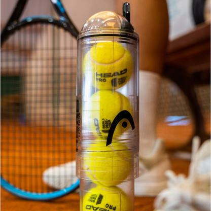 Clear container with tennis balls and a racket in the background
